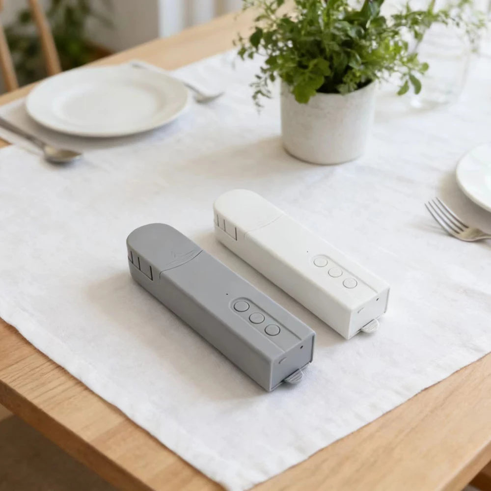Two electronic devices on a white tablecloth with a plant and plates in the background
